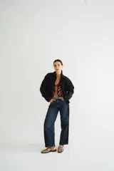 Campera de jean negra tipo bomber con cuello redondo y cierre frontal con botones metálicos. Presenta un parche rectangular con diseño gráfico en la espalda.