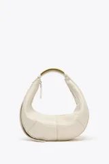 Bolso ovalado color crudo con asa de hombro metálica dorada. Tiene pespuntes en contraste, bolsillo interior con cremallera y cierre principal con cremallera y tirador.