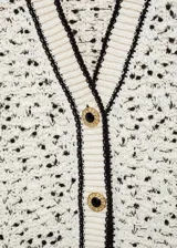Cárdigan de punto grueso color crema con motas negras, cuello pico, cierre frontal con botones efecto joya, dos bolsillos de parche delanteros y ribete en contraste color negro.