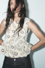 Top sin mangas color crudo con estampado de flores negras, cuello redondo, peplum en la cintura y cierre trasero con cremallera metálica.