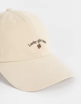 Gorra color beige con bordado frontal de la frase "Lucky Girl Club" y un trébol de cuatro hojas.