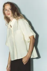 Camisa color crema de cuello solapa y manga corta con vuelta. Tiene un bolsillo de parche en el delantero, bajo asimétrico con aberturas laterales y cierre frontal con botones.