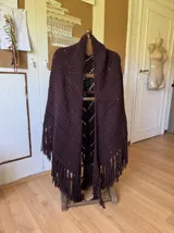 Ruana tejida a mano en color lila, confeccionada en lana merino. Presenta un diseño de punto con calados diagonales y terminación de flecos largos en el ruedo.