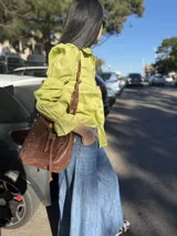 Bolso tipo bucket de cuero color marrón con tachas plateadas. Tiene correa larga regulable para usar al hombro y cierre con cordón ajustable.