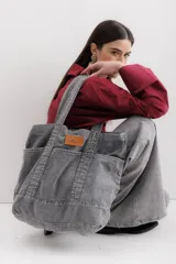 Bolso tote grande de jean gris con asas de mano y asa larga para colgar. Se cierra con un botón de metal a presión y contiene tres bolsillos exteriores.