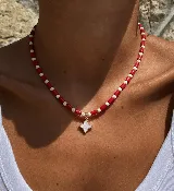 Gargantilla corta con cuentas tubulares rojas y cuentas redondas blancas intercaladas. Dije central en forma de estrella de cuatro puntas con borde dorado y centro de nácar.