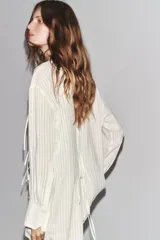 Camisa blanca a rayas verticales, de cuello redondo y manga larga con puños. Presenta cierre frontal con cordones ajustables con lazadas y abertura en la espalda ajustable con lazadas.
