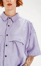 Camisa de manga corta color lavanda con doble bolsillo y ojal delantero con solapa en uno de ellos. Tiene calce oversize y está realizada en tejido plano suave al tacto.