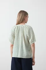 Camisa de viscosa con estampado de pequeños lunares verdes sobre fondo blanco o crema. Presenta escote en V con botones frontales y mangas cortas abullonadas con elástico en el puño.