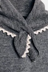Chaqueta de punto gris oscuro, corte cropped y manga larga, con cuello redondo que incorpora una bandana anudada al frente. Los bordes del cuello, puños y ruedo están terminados con una puntilla de encaje color beige claro. Cierre frontal con botones oscuros.