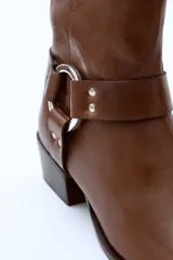 Bota de cuero negra con caña media, puntera ligeramente afilada y detalle de correa con herraje metálico circular y tachas. Taco bajo y ancho.