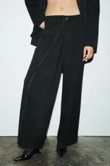Pantalón negro de tiro medio con trabillas, falso bolsillo de vivo en espalda, detalle de pespuntes combinados a contraste y pinzas. Cierre frontal con cremallera y botón.