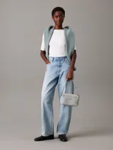 Cartera tipo camera bag de color blanco/gris claro, confeccionada en piel sintética con textura de monograma en el bolsillo frontal. Cuenta con dos correas desmontables: una ancha y otra fina, ambas ajustables. El bolsillo frontal tiene el logo de Calvin Klein en metal plateado.