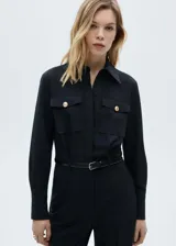 Mono largo negro de corte slim, con cuello camisero, cierre frontal abotonado, cinturón con hebilla dorada, dos bolsillos de parche con solapa en el pecho y puños abotonados.