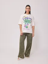Remera color crudo de corte oversized con estampado de palmeras, flores y texto 'California' y 'Rusty Tribe'.