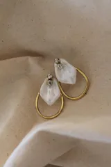 Aros colgantes dorados con forma de aro abierto, de los cuales cuelga una pieza irregular de nácar o resina blanca iridiscente. En la parte superior, donde se enganchan al lóbulo, tienen dos pequeños cristales brillantes.