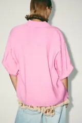 Camisa rosa de cuello solapa y manga corta con vuelta. Bajo asimétrico con aberturas laterales. Cierre frontal con botones ocultos por solapa.