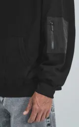 Hoodie negro de corte oversize con capucha, bolsillo tipo canguro y detalle de bolsillo con cierre en la manga izquierda.