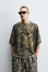 Sudadera relaxed fit con estampado de hojas en tonos verde, marrón y negro. Tiene cuello redondo, manga corta y bajo irregular con aberturas laterales.