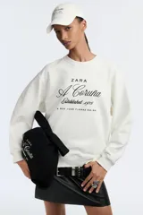 Sudadera negra de cuello redondo y manga larga, con bordado blanco en el frente que dice 'Zara A Coruña Established 1975 Rúa Juan Flórez 64-66' y un logo pequeño con el número 50 en la parte superior.