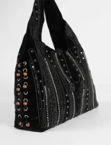 Bolso de mano tipo hobo o baguette, confeccionado en terciopelo negro. Presenta un diseño ornamentado con apliques de mostacillas blancas y negras formando líneas verticales y motivos florales. El lateral tiene ojales metálicos por donde pasan cordones negros, adornados con cuentas de madera redondas.