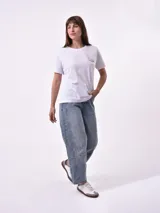 Remera blanca de algodón con cuello redondo y manga corta. Presenta un pequeño bordado en el pecho izquierdo con una figura que parece ser una persona acostada en una tabla de surf o flotador.