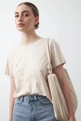 Remera de algodón color beige claro con cuello redondo y manga corta. Presenta un bordado en relieve de tres flores en el frente.