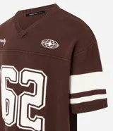 Remera oversized deportiva color marrón con cuello en V, rayas blancas en las mangas y en la parte inferior, y el número "62" estampado en blanco en el frente. Incluye el bordado "Humanity" y un logo de globo terráqueo.