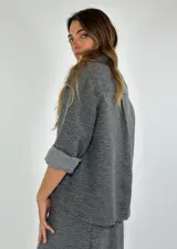Chaqueta color gris con textura, de corte recto, cuello camisero, cierre frontal con botones, mangas 3/4 con puños arremangados, dos bolsillos de parche en el pecho y ruedo recto.