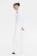 Camisa blanca de algodón con cuello clásico, abotonada adelante y corte holgado. Tiene mangas largas con puños sencillos y bajo redondeado con espalda algo más larga.