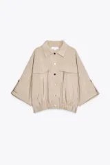Camisa color beige de cuello solapa y manga corta con trabilla y botón. Tiene bolsillos delanteros de plastrón con solapa, bajo con elástico y cierre frontal con botones a presión.