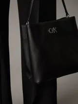 Cartera tipo saco Calvin Klein color negro, con correa ajustable para el hombro, compartimento central con cremallera, compartimento con cierre giratorio en la parte delantera, compartimento magnético en la parte trasera y herrajes plateados con el monograma de la marca.