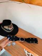 Cinturón ancho de doble capa, con capa superior de gamuza negra o marrón y capa inferior de cuero color camel. Presenta múltiples ojales metálicos y hebilla ovalada metálica de color dorado.