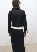 Chaqueta corta de cuero negro con cuello camisero, cierre frontal con botones a presión plateados, bolsillos laterales y puños abotonados.