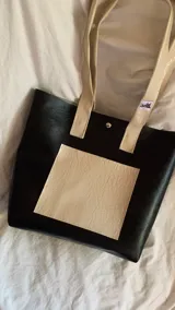 Bolso tipo tote bag negro con bolsillo rectangular color crema en el frente y asas cortas color crema.