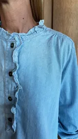 Blusa blanca con textura de lunares, cuello con volados, cierre parcial con botones y mangas largas con puños abotonados.