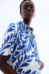 Camisa de manga corta con estampado de hojas y flores en tonos azules sobre fondo blanco. Tiene cuello clásico, botones frontales, corte holgado y canesú con pliegue en la espalda.