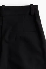Pantalón sastrero negro de corte holgado con tiro alto y cierre con corchete y botón ocultos. Piernas amplias con pinzas, pliegues adelante y atrás, bolsillos al bies y bolsillos decorativos atrás.
