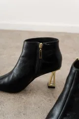 Botín de cuero negro con punta fina y cierre lateral. Taco dorado con diseño geométrico.