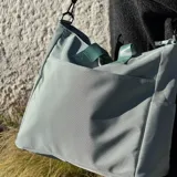 Bolso tipo tote color gris claro, con doble asa de mano y correa larga regulable. Tiene dos bolsillos laterales y un bolsillo frontal.