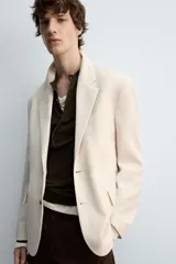 Blazer color beige confeccionado en tejido con lana, de corte relajado. Tiene cuello con solapas de muesca, manga larga con puños abotonados, bolsillo de vivo en el pecho, bolsillos de solapa en la cadera, bolsillo interior, bajo con abertura en el centro de la espalda y cierre frontal con botones.