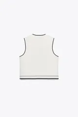 Chaleco de punto color blanco con cuello redondo y manga sisa. Tiene falsos bolsillos de vivo en la parte delantera y detalle de bandas combinadas a contraste en color negro. Cierre frontal con botones a presión.