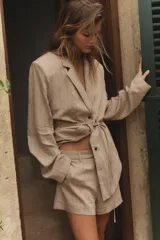 Blazer color beige con finas rayas verticales negras, cuello solapa, manga larga con hombreras, cinturón tipo cordón en el mismo tejido y cierre frontal con botones.