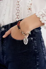 Pulsera de cuentas de piedras semipreciosas en tonos rojizos, negros y verde claro, con cierre de mosquetón dorado y un dije colgante en forma de escudo plateado con una cruz en relieve.