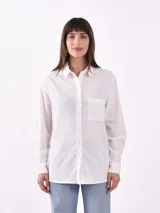 Camisa blanca de manga larga, con cuello clásico, botones frontales y un bolsillo de parche en el pecho izquierdo.