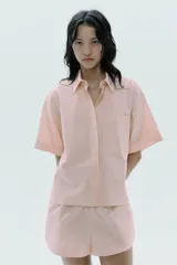 Camisa color rosa claro a rayas verticales finas color marrón, de popelín, con cuello solapa y manga corta con dobladillo. Tiene un bolsillo de parche en el pecho, aberturas laterales en el bajo y pinzas en la espalda. El cierre frontal es con botones ocultos.