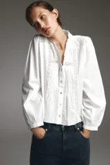 Camisa blanca de cuello subido y manga larga, con detalle de entredós combinados a tono y cierre frontal con botones.