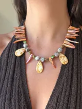 Collar corto con cuentas de cristal checo, piedras facetadas y colgantes en forma de lágrima de nácar teñido en tonos crema y naranja. Incluye detalles en forma de colmillo teñido en tonos naranjas y cuentas redondas con textura agrietada en tonos grises y verdes.