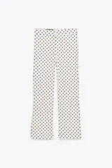 Pantalón largo de tiro alto con estampado de lunares negros sobre fondo blanco. Tiene falsos bolsillos de vivo en la espalda, detalle de solapa en el delantero y bajo acampanado. Se cierra con cremallera y gancho metálico.
