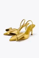 Zapato de tacón destalonado color dorado, con efecto satinado, punta fina, tira trasera elástica y maxi lazo decorativo en la parte delantera.